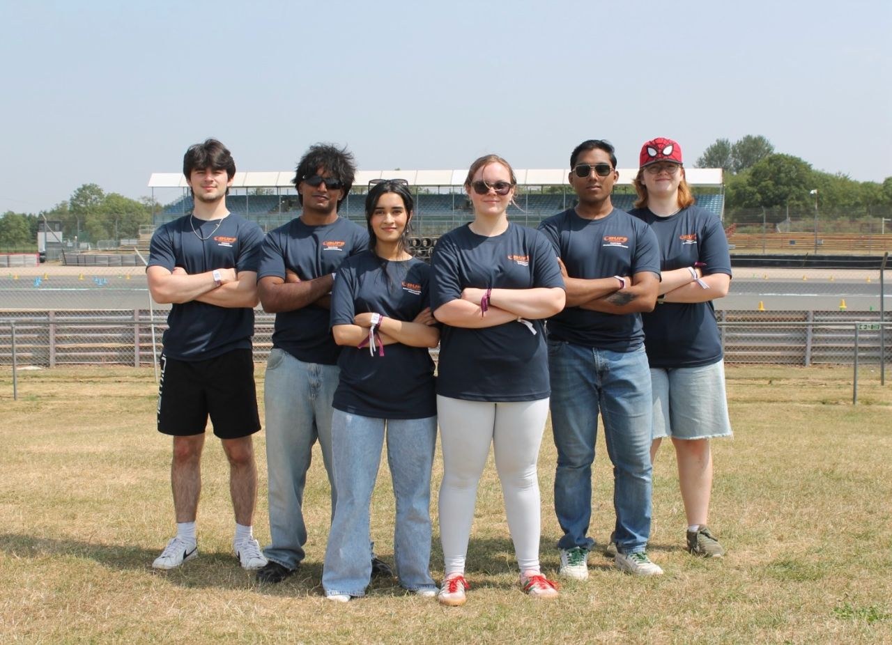 The Bournemouth Formula Student team, six members, standing arms-crossed on the grass at Silverstone with the grandstand and track behind them